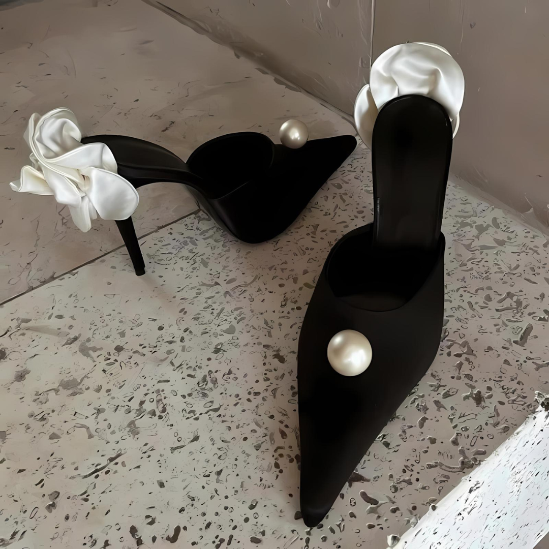 Pearl Blossom Heels auf einer Treppe. Die eleganten Stilettos mit spitzer Zehenpartie, Satin-Obermaterial und zarten Perlen- sowie Blumenapplikationen präsentieren sich mit einem Slingback-Riemen und hohem Absatz für einen glamourösen Look.