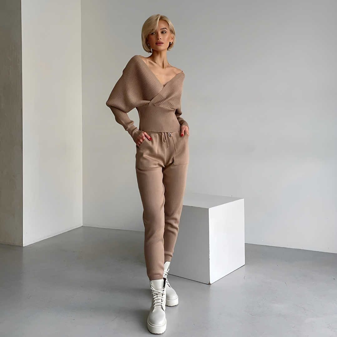 Frau in elegantem, schulterfreiem Pullover und passender Hose des Sets Emily in Vorderansicht. Das zweiteilige Set aus weichem Strickstoff bietet Komfort und Wärme und kombiniert einen sportlichen Look mit einem femininen Touch. Ideal für Freizeitaktivitäten und entspannte Tage.“