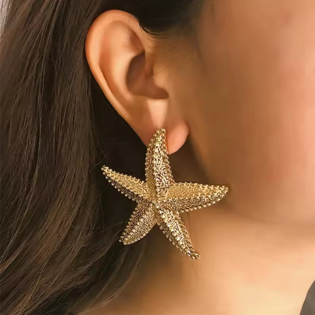 Goldene Ohrstecker im Seestern-Design, getragen von einer Frau. Die auffälligen Ohrringe kombinieren maritime Eleganz mit luxuriösem Glanz und verleihen jedem Outfit einen glamourösen Touch.