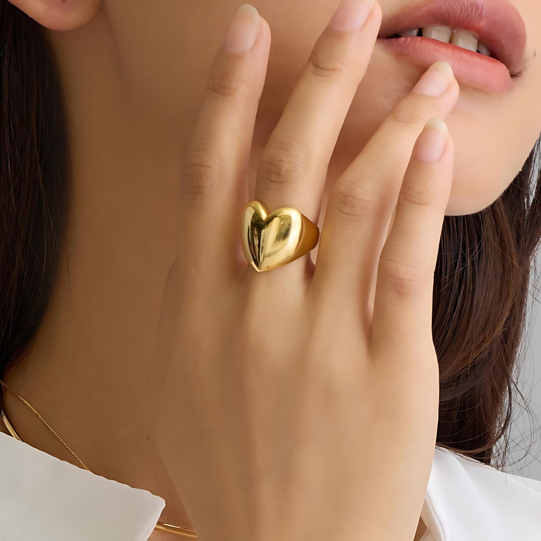 Goldener Herz-Ring, getragen von einer Frau, die sich an die Wange fasst, im Licht glänzend. Der Ring aus hochwertigem goldfarbenem Metall betont die luxuriöse Textur und elegante Ausstrahlung des Schmuckstücks. Ideal für romantische Anlässe oder als stilvolles Accessoire für den Alltag.