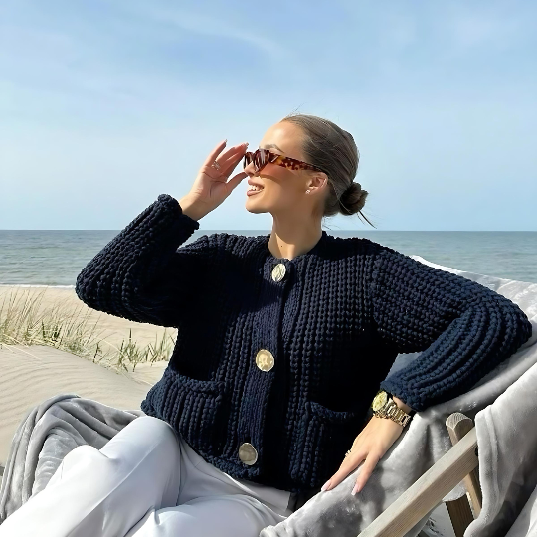 Sitzende Frau in elegantem Cardigan Ava, bestehend aus hochwertigem, weichem Strickmaterial mit klassischem O-Ausschnitt und Knopfleiste, in Vorderansicht. Der Cardigan hat lange Ärmel und eine lockere, entspannte Passform, die maximalen Komfort und Wärme an kühleren Tagen bietet. Ideal für Alltag, Büro-Outfits und entspannte Wochenenden im Herbst und Winter.