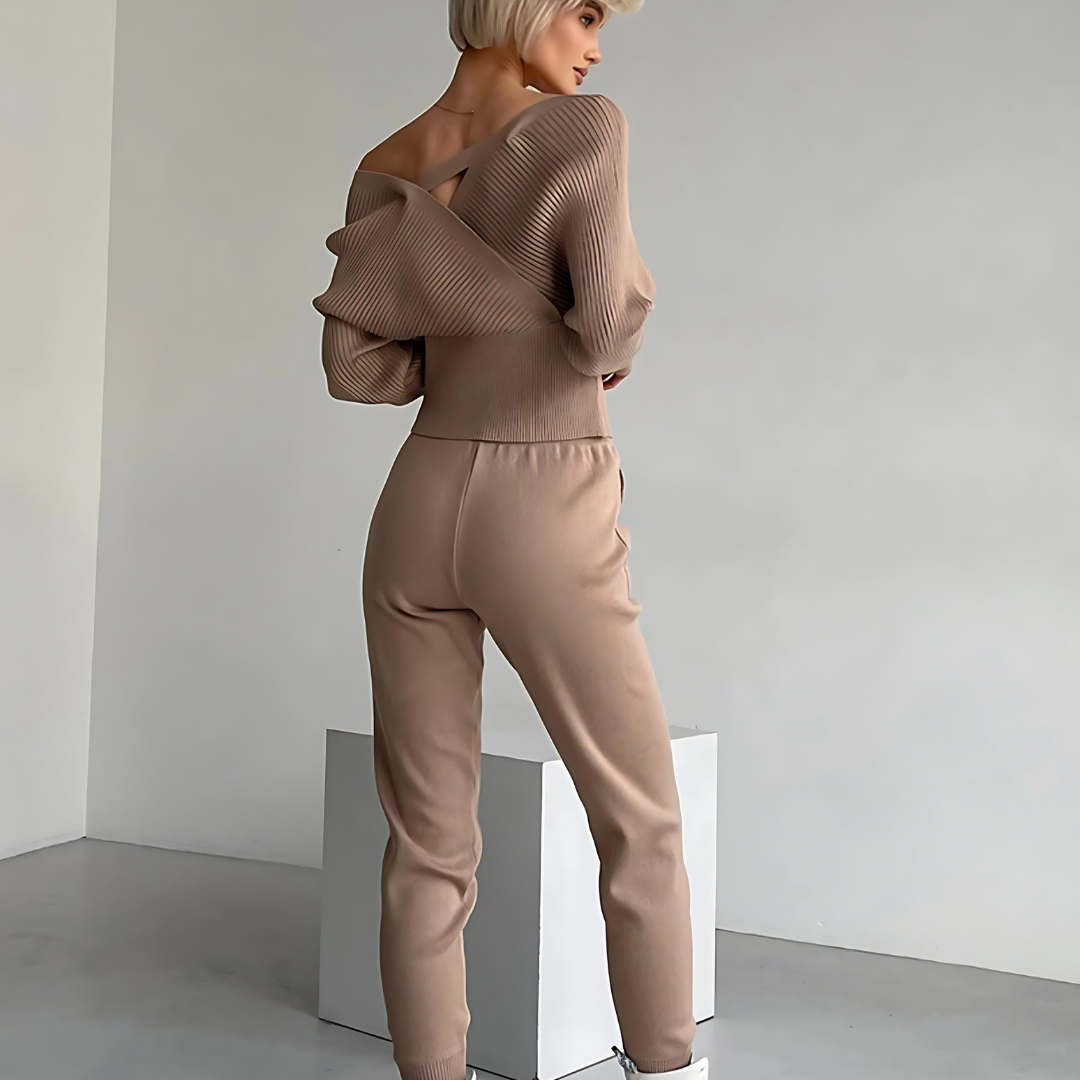 Rückansicht des Sets Emily, getragen von einer Frau. Das zweiteilige Set besteht aus einem schulterfreien Pullover und einer passenden Hose aus weichem Strickstoff, das Komfort und Wärme bietet. Der moderne und sportliche Look ist ideal für entspannte Tage und stilvolle Freizeitoutfits.