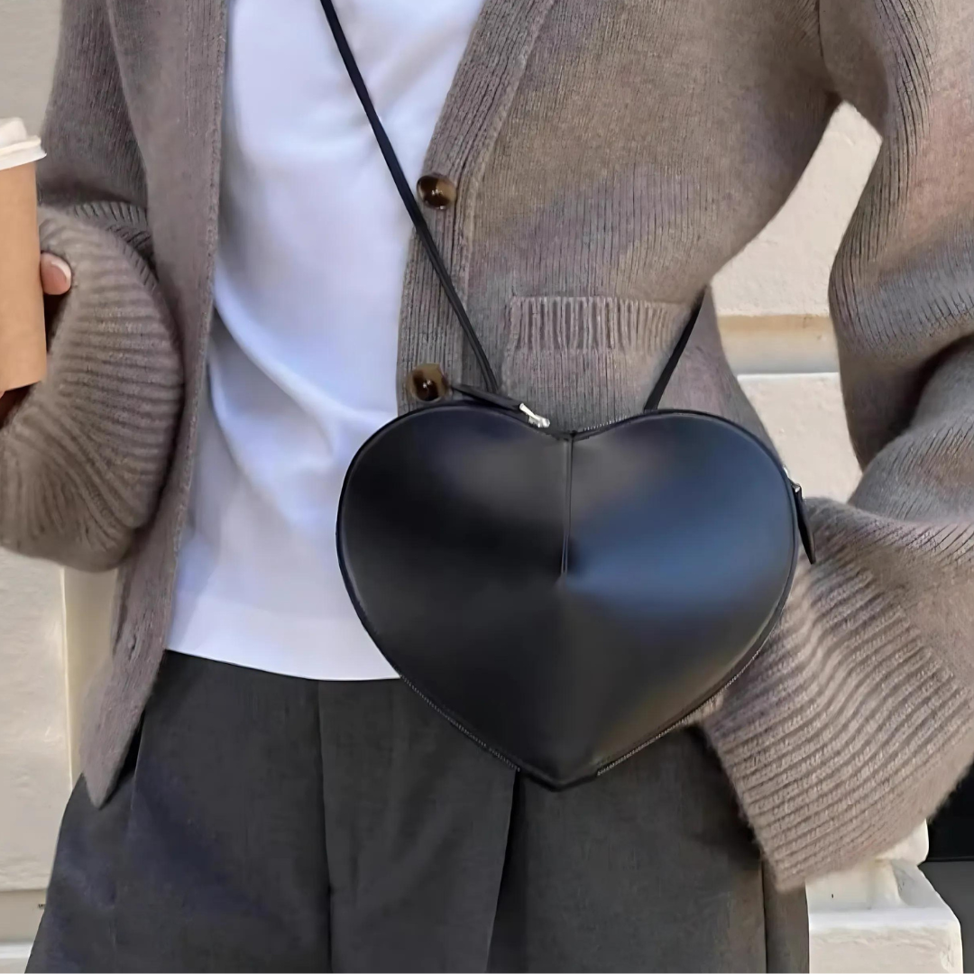 Frau trägt die schwarze Herz-Tasche umgehängt über der Schulter. Die herzförmige Silhouette der Tasche ist deutlich sichtbar und verleiht dem Outfit der Frau eine elegante, trendige Note. Ideal für modebewusste Frauen, die ein stilvolles und praktisches Accessoire suchen.
