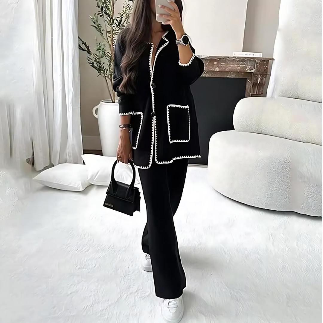 Frau in elegantem Set Elena in klassischem Schwarz, bestehend aus einer einreihigen Strickjacke mit langen Ärmeln und Knopfverschluss sowie einer passenden Hose mit elastischem Bund und geradem Schnitt, in Vorderansicht. Das Set verfügt über weiße Applikationen an den Nähten und Taschen, die einen stilvollen Akzent setzen. Das hochwertige, weiche Strickmaterial sorgt für Komfort und eine entspannte Silhouette, ideal für Alltag, Büro, Freizeit oder besondere Anlässe.