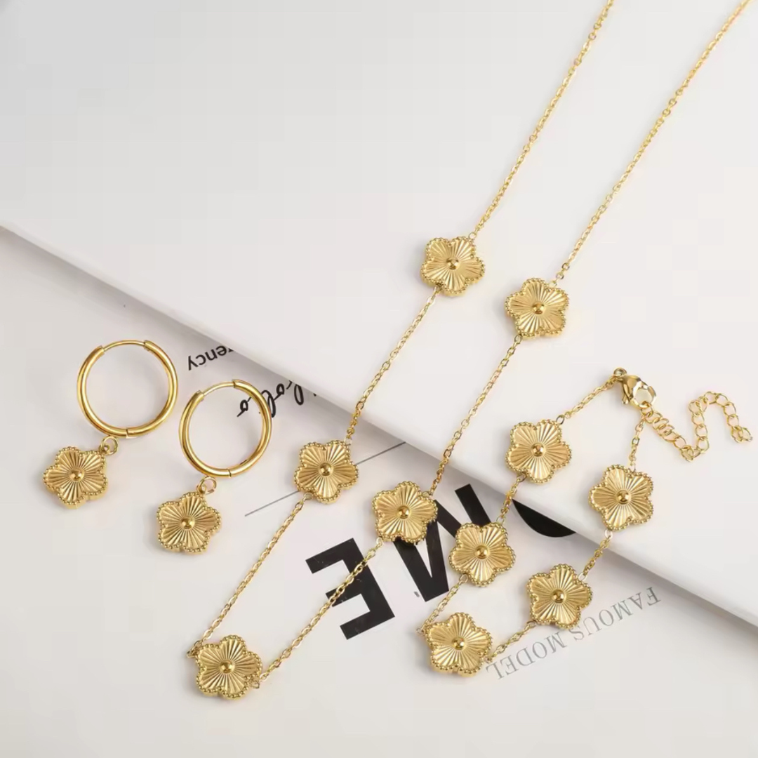 Elegantes Schmuckset Cloverleaf bestehend aus goldfarbenen Ohrringen, Halskette und Armband mit filigranem Kleeblatt-Anhänger auf neutralem Hintergrund. Das Set aus hochwertiger, glänzender Legierung symbolisiert Glück und ist vielseitig für verschiedene Anlässe.